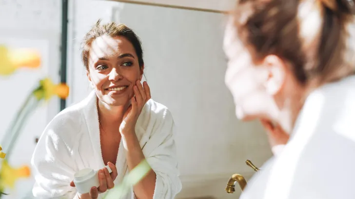 Beauty portrait of young smiling woman with dark long hair in white bathrobe put day nourishing moisturizer cream on clean fresh skin face and hands near mirror in bathroom at home