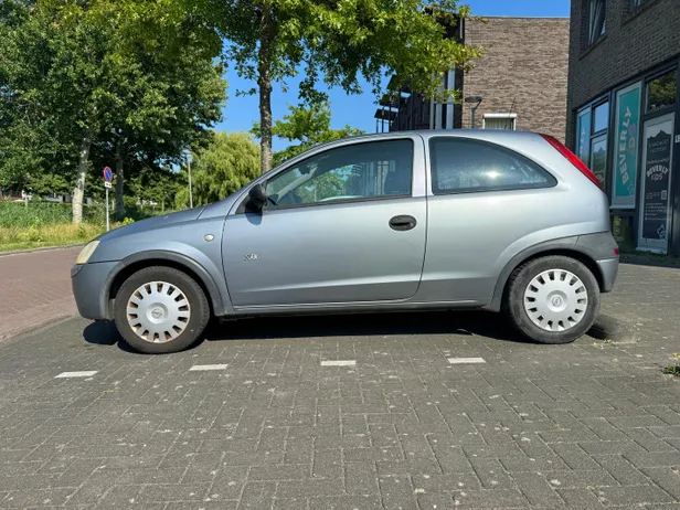 Oudere grijze Opel Corsa geparkeerd op een klinkerbestrating langs een Nederlandse straat.