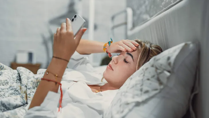 Top view unhappy woman feeling headache after sudden awakening by phone call, message signal or alarm in early morning, exhausted young female suffering from insomnia or migraine, lying in bed.