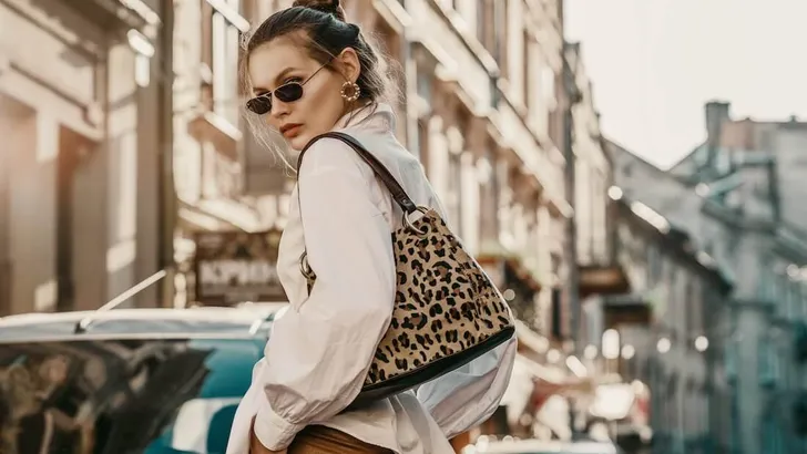 Outdoor autumn fashion portrait of elegant, luxury woman wearing sunglasses, trendy white shirt, leather trousers, with animal, leopard print bag, walking in street of European city. Copy, empty space