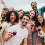 Group of young adult best friends having fun toasting a red wine glasses at rooftop reunion or birthday party, drinking alcohol. Happy people enjoying on a social gathering celebrating together