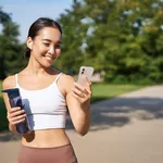 Fitness woman with water bottle and smartphone, jogging in park and smiling, looking at her mobile phone app, checking sport application