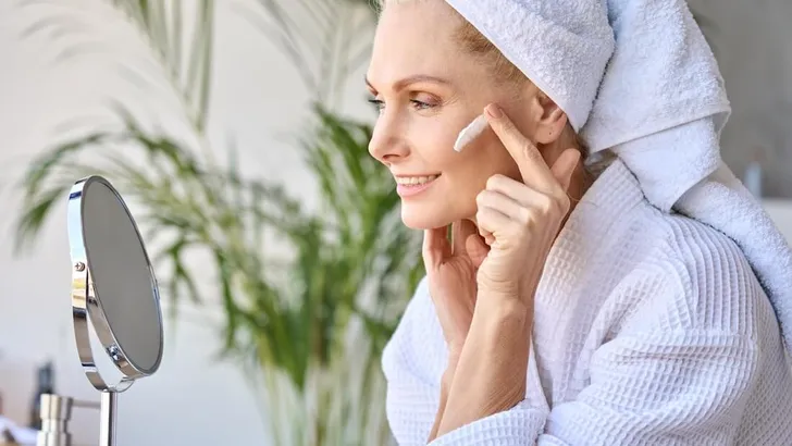 Smiling beautiful older lady looking at mirror applying cream. Skincare concept.