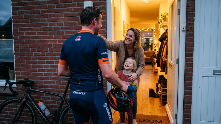 Een mannelijke wielrenner wordt na zijn fietstocht in de deuropening van zijn huis verwelkomd door zijn partner en kind.