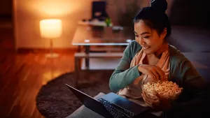 Happy Asian woman eats popcorn while watching movie on laptop at night at home.