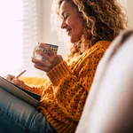 Happy cute lady at home write notes on a diary while drink a cup of tea and rest and relax taking a break. autumn colors and people enjoying home lifestyle writing messages or lists. Blonde curly beautiful lady sit down in the house