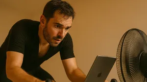 Close-up van een zwetende wielrenner op een indoortrainer, met een ventilator en tablet naast het stuur in warm licht.