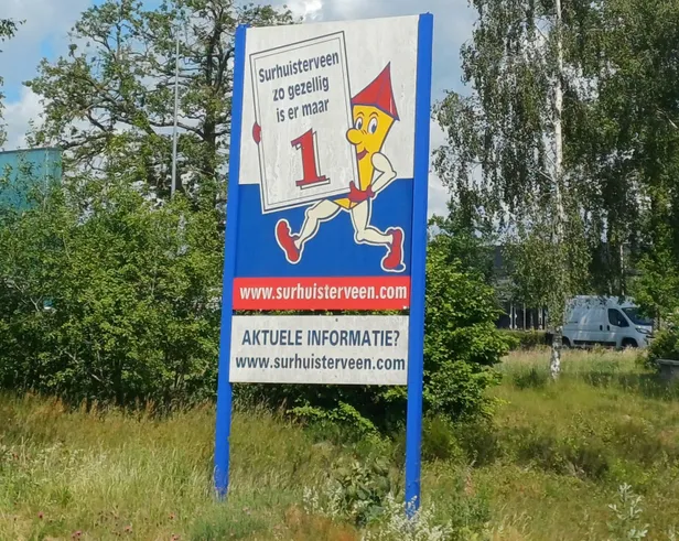 Bijzondere citymarketing in Surhuisterveen met een blauw bord en een gespierde, antropomorfe kerktoren met armen en benen langs de weg.