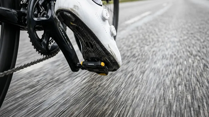 Een close-up van moderne wielerschoenen met draaisluitingen, strak ingeklikt in de pedalen van een racefiets die op hoge snelheid over het asfalt rijdt.