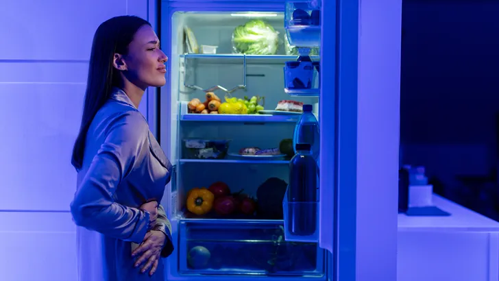 Een vrouw opent de koelkastdeuren en zoekt naar snacks, voelt honger en raakt haar buik aan.