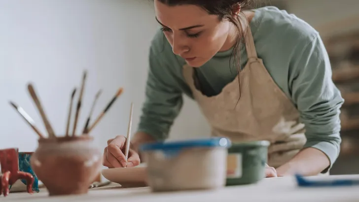 Skillful young woman in apron painting pottery at workshop