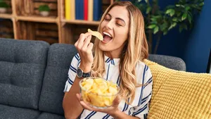 Young blonde woman eating chips potatoes sitting on sofa at home