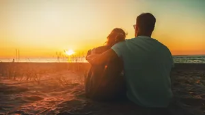 young lovers couple sitting in sand on beach at romantic golden sunset. back view