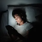 Young woman using her cellphone while lying in bed at night