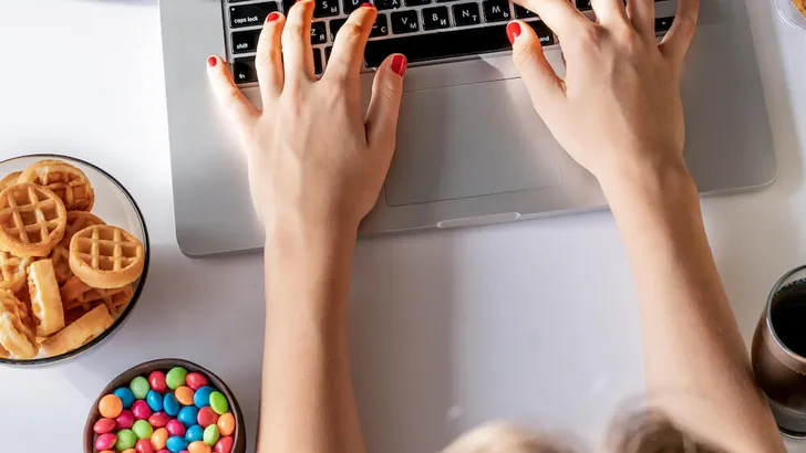 Girl works at a computer and eats fast food. Unhealthy food: chips, crackers, candy, waffles, cola. Junk food, concept.