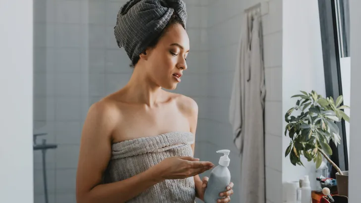 Young African American Woman Wrapped in a Towel after a Shower Holding a Skin Care Product
