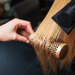 Female client in beauty salon. Close-up of hairdressers hand drying blond hair with hair dryer and round brush, doing new hairstyle