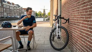 Een moderne witte racefiets geparkeerd met een slimme truc: het stuur is naar binnen gedraaid tegen een klassieke bakstenen muur op een Nederlands terras, waardoor hij muurvast staat zonder standaard. Een wielrenner staat ernaast met koffie.