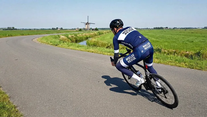 Mannelijke wielrenner met helm, armstukken en beenstukken snijdt geconcentreerd een bocht aan op een vlakke Nederlandse weg