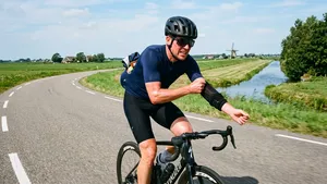 Een zwetende wielrenner met fietshelm probeert al fietsend krampachtig zijn warme armstukken uit te trekken.