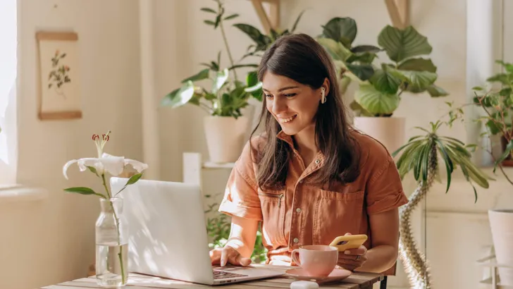 Positive young mixed race woman using a laptop and smartphone at home.Cozy home interior with indoor plants.Remote work, business,freelance,online shopping,e-learning,urban jungle concept