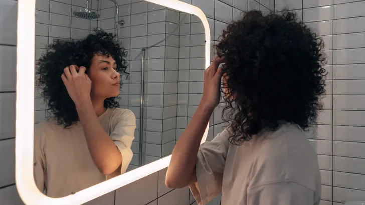 Young pretty woman checking herself in the mirror in modern bathroom. Putting curly hair behind ear
