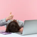 I am fine. Extremely tired sleepy woman lying on table with laptop and cup of coffee showing thumbs up, exhausted with overwork, vitamin deficiency