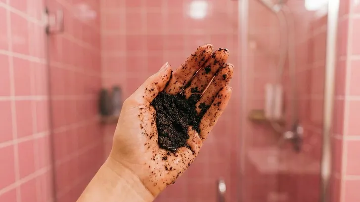 Close-up van een vrouwenhand gevuld met koffiescrub, tegen een achtergrond van roze badkamertegels.