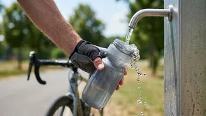 Close-up van een fietser die een transparante bidon vult met stromend water bij een openbaar watertappunt in een zonnig park.