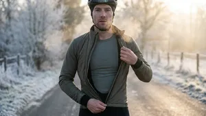 Een mannelijke wielrenner staat op een bevroren weg in een winters landschap en ritst zijn olijfgroene winterjack open, waardoor een technisch thermoshirt zichtbaar wordt. Zijn adem vormt wolkjes in de koude lucht.