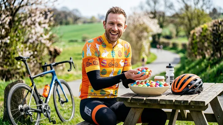 Een vrolijke wielrenner in een speciaal paaswielershirt met arm- en beenstukken zit aan een buitentafel na een rit. Hij glimlacht naar een grote schaal met kleurrijke chocolade paaseitjes, met zijn helm rustend op de tafel naast hem.
