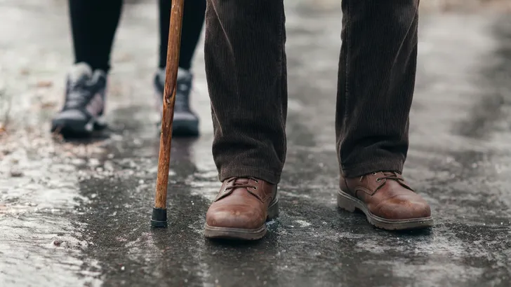 Een persoon, van wie alleen de benen en een hand zichtbaar zijn, draagt een grijze broek en bruine schoenen en houdt een houten wandelstok vast in de gladheid.