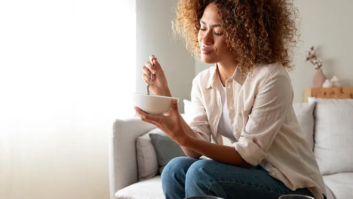 Young relaxed pensive African American woman eating healthy breakfast sitting on the couch. Copy space.