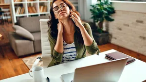 Tired business woman in stress works at a laptop while sitting at a table at home and talking on the phone. Freelance, work from home.