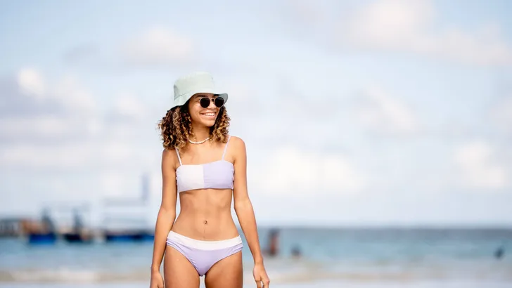 Smiling young woman in a sun hat and bikini walking on a beach