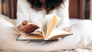 Unrecognizable young woman turning the pages of a book while reading in bed covered with the duvet