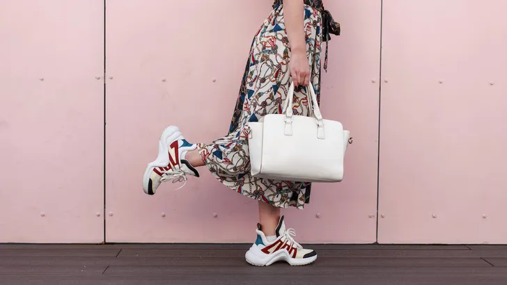 Stylish woman with a white bag in a vintage dress with fashionable sneakers posing near the pink wall. Close-up. The concept of shopping