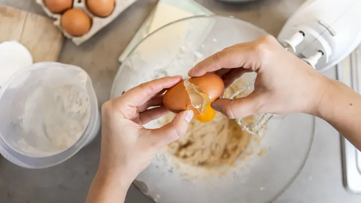 Vrouw breekt rauw ei open boven mengschaal voor het maken van een cake.