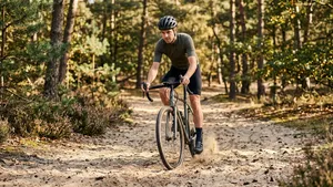Een mannelijke wielrenner op een gravelbike rijdt met een gecontroleerde techniek en gefocuste blik door een zandstrook op een bospad. Hij draagt een helm en zijn gewicht is naar achteren verplaatst.
