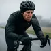 Wielrenner in zwarte winterkleding fietst door grijze, natte kou, met rode wangen en zichtbare adem terwijl hij tegen de wind in leunt.