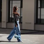 Full length beautiful young brunette woman Wearing black leather jacket and wide blue jeans, sneakers, sunglasses and small handbag with chain, walking street on sunny day