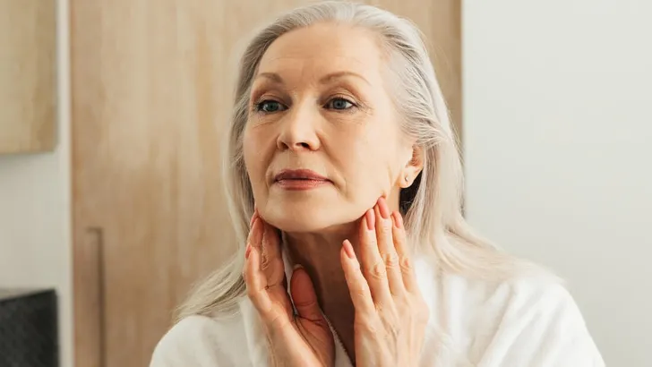 Senior woman stretching skin on her face in front of a mirror