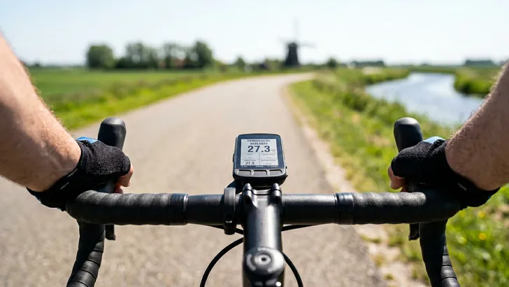Een mannelijke wielrenner kijkt naar zijn fietscomputer die prominent 'GEMIDDELDE SNELHEID 27.3 km/u' aangeeft, terwijl hij over een zonnige dijk fietst.