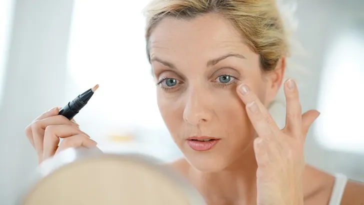 Middle-aged blond woman putting eye concealer in front of mirror