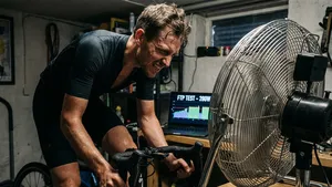 Een mannelijke wielrenner die tot het uiterste gaat tijdens een FTP-test op een indoortrainer in een donkere garage, met een grote ventilator voor zich en een laptop die 'FTP TEST - 280W' weergeeft.
