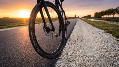 Close-up van het voorwiel van een all-road racefiets die op de grens van een asfaltweg en een gravelpad rijdt tijdens zonsondergang.