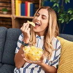 Young blonde woman eating chips potatoes sitting on sofa at home