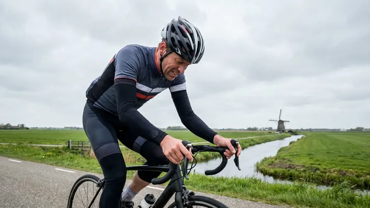 Een mannelijke wielrenner met arm- en beenstukken fietst vermoeid en zwoegend op een vlakke polderweg in Nederland, met een molen in de achtergrond.