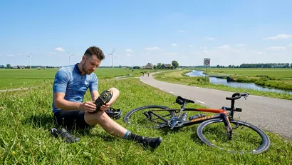Een mannelijke wielrenner zit in de berm zonder helm en inspecteert nauwkeurig het schoenplaatje onder zijn fietsschoen.