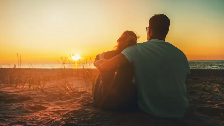 young lovers couple sitting in sand on beach at romantic golden sunset. back view